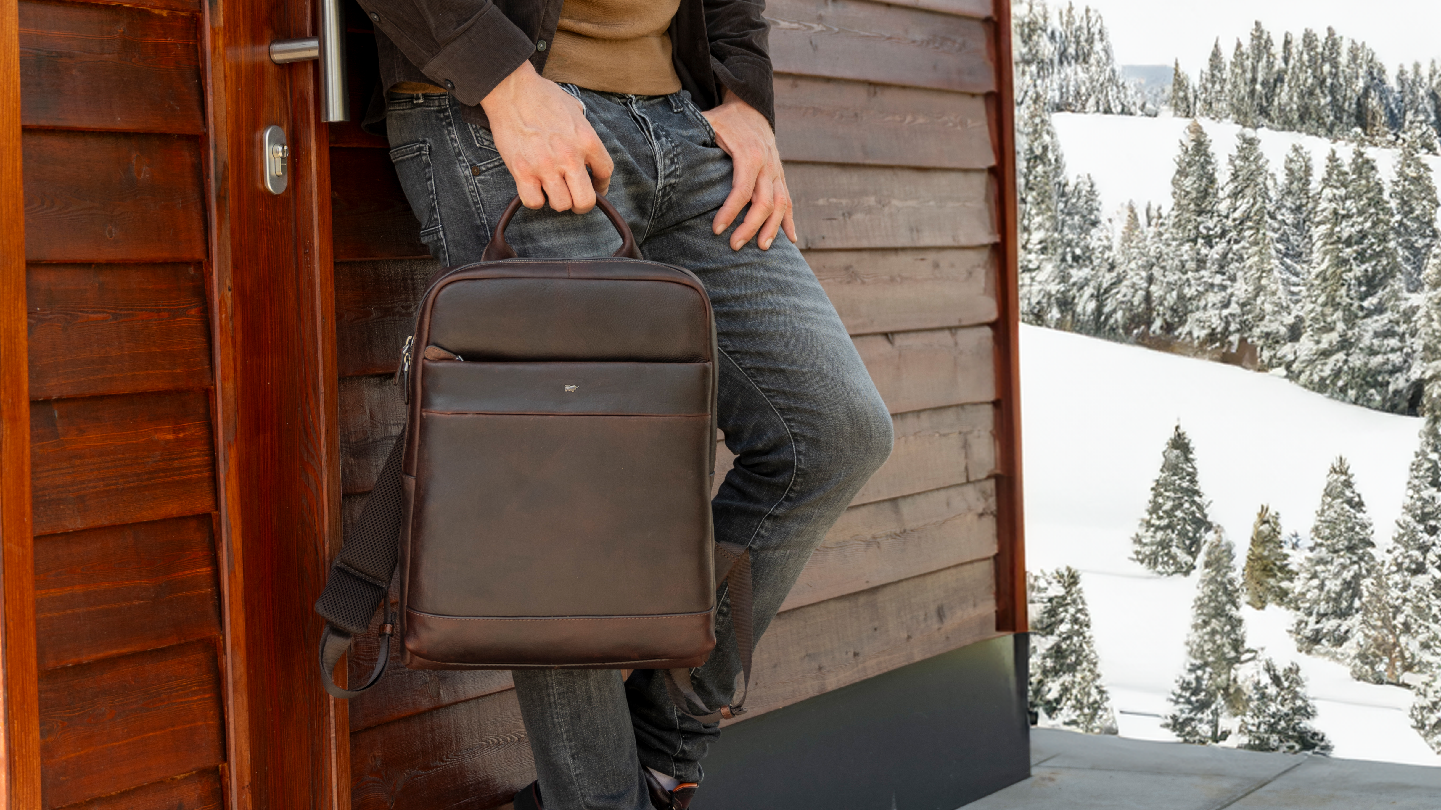 Mann steht mit Lederrucksack in einer winterlichen Landschaft mit Schnee